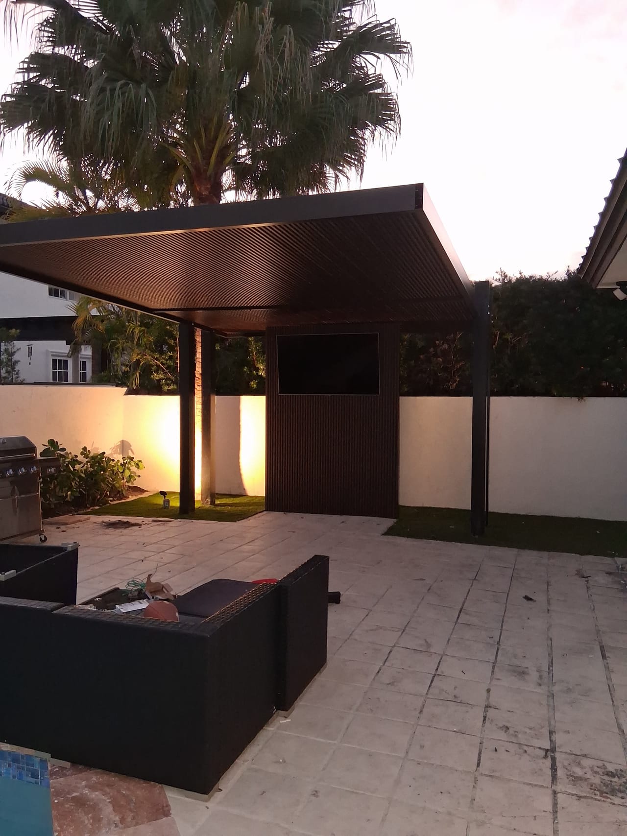 Poolside cabana at dusk with a dark slat wall, recessed television niche and warm uplighting washing the courtyard wall behind it