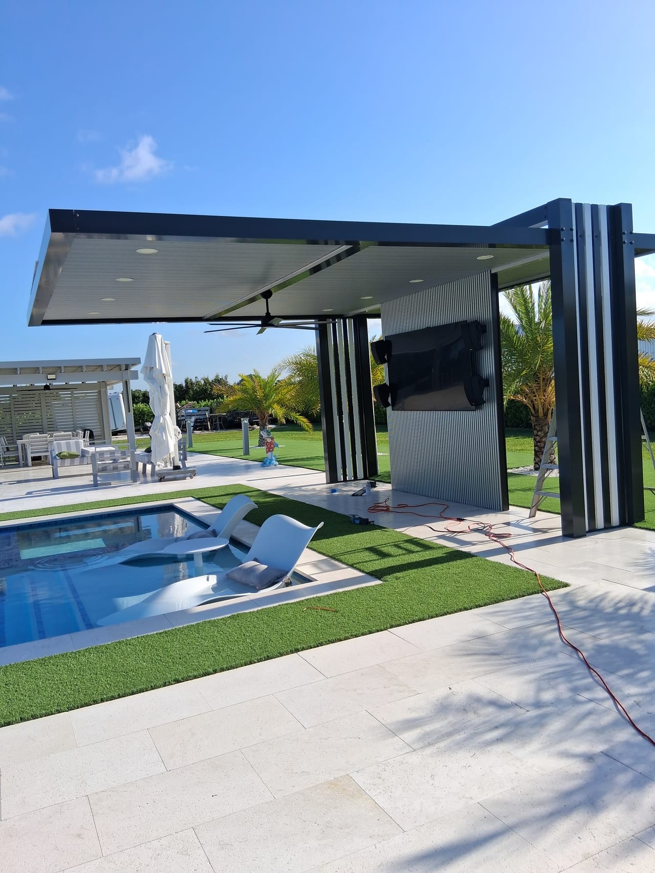 Freestanding aluminum pergola with a cantilevered roof and a fluted media wall holding an outdoor television beside a pool