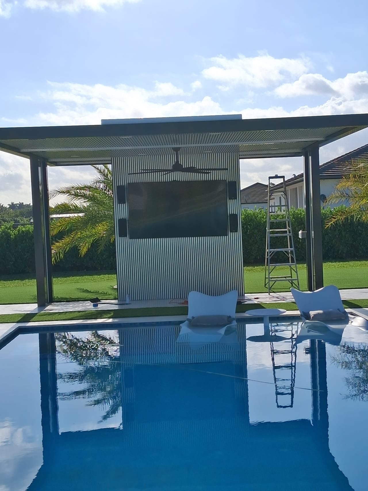 Modern pergola framing a pale fluted wall with a wall-mounted outdoor television and ceiling fan, reflected in a swimming pool