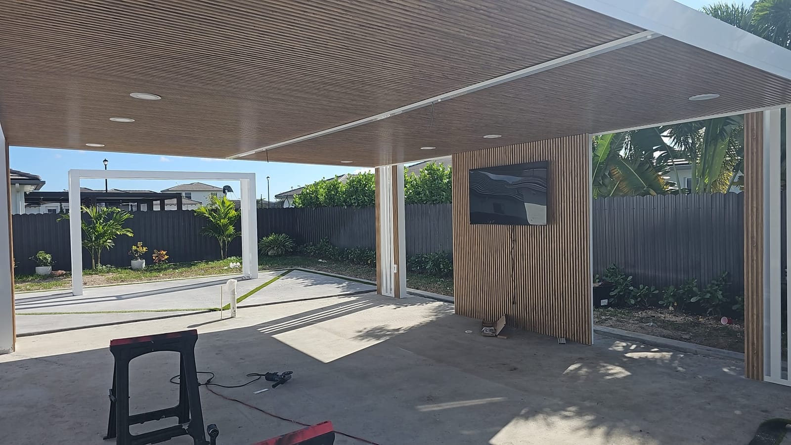 Covered patio with a warm wood-look soffit and slat feature wall, looking out through a white pergola frame onto a South Florida garden