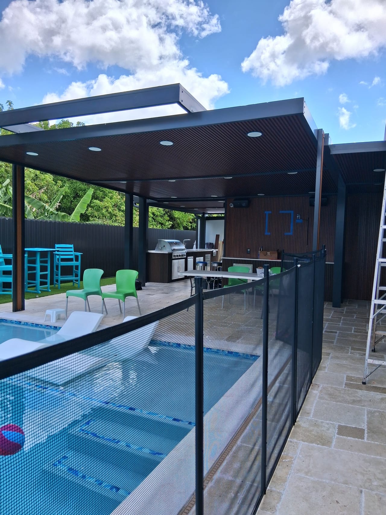 Dark aluminum pergola with a wood-look soffit covering an outdoor kitchen and bar beside a travertine pool deck