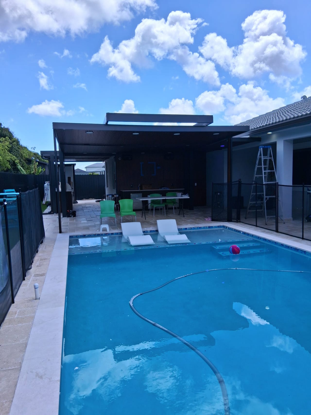 Poolside view of an aluminum pergola sheltering an outdoor kitchen, dining table and seating on a South Florida patio
