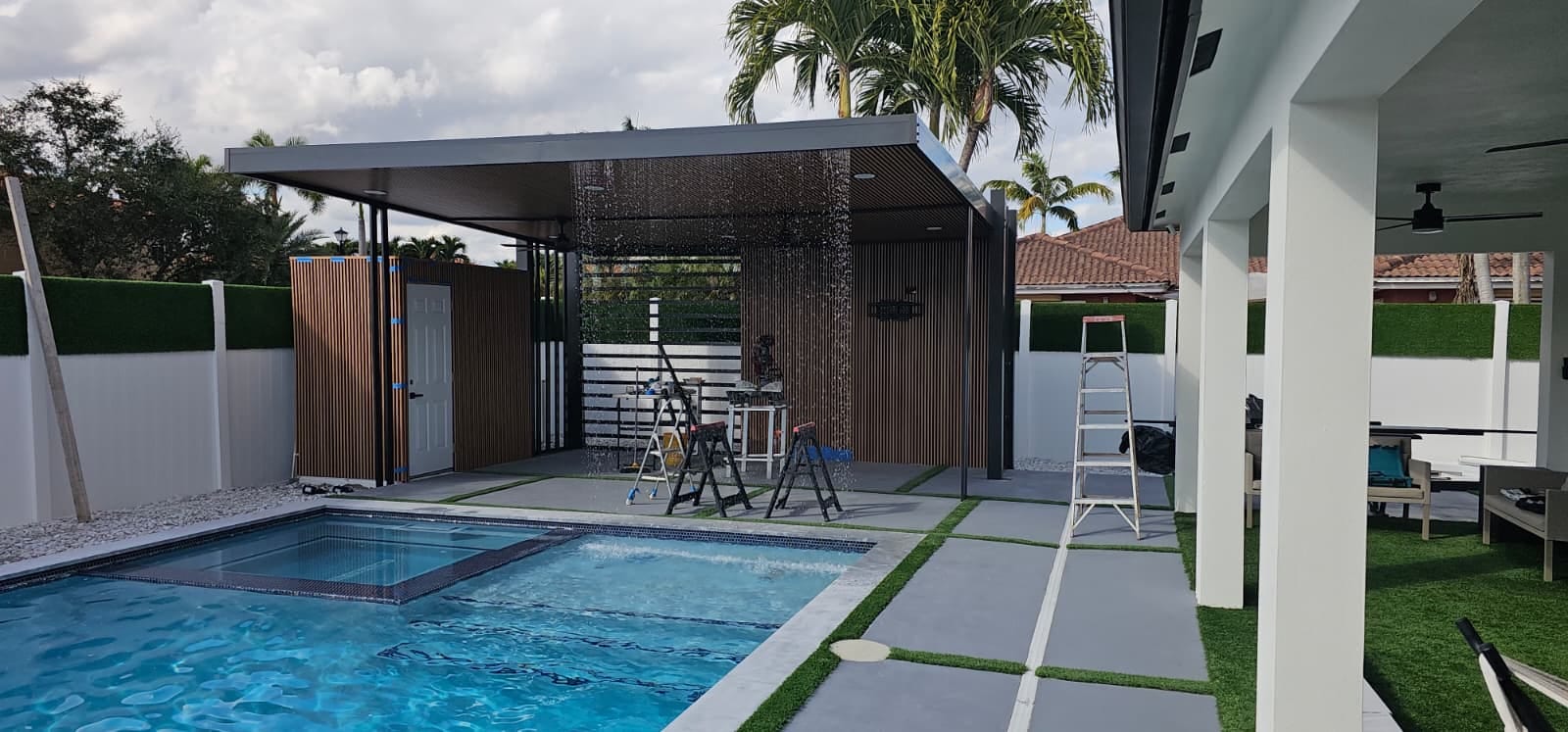 Bronze aluminum pool cabana with slatted privacy walls and a rain-curtain water feature falling beside a South Florida swimming pool