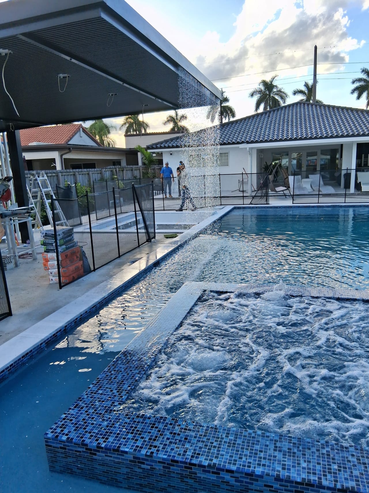 Rain curtain falling from the edge of a modern pool cabana roof, seen across a pool with a bubbling spa in the foreground