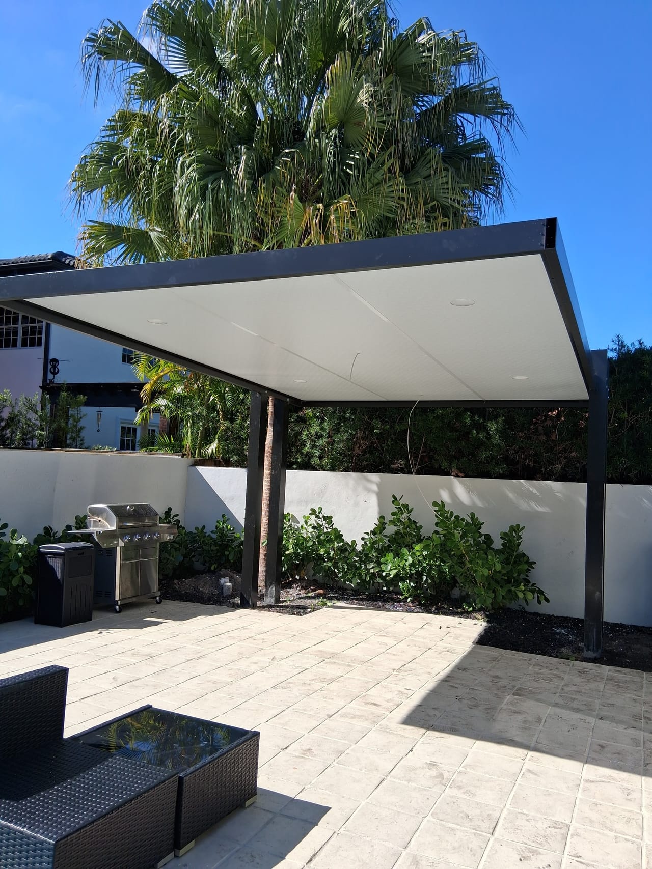 Small white solid-roof pergola on a stamped concrete patio shading a grill and lounge seating against a white garden wall